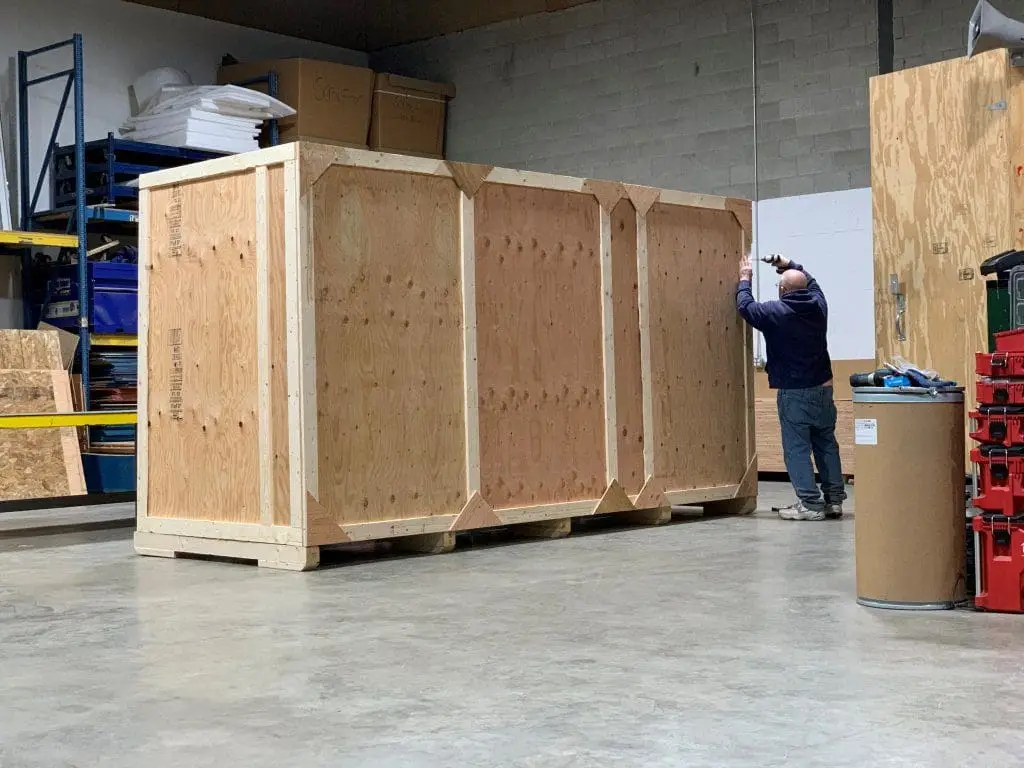 Person standing beside a large wooden shipping crate in a warehouse.