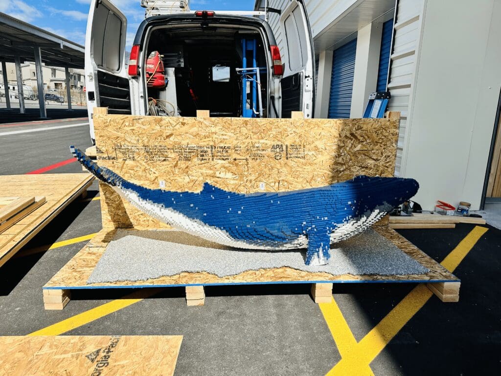 Lifelike sculpture of a whale made from wooden blocks, displayed on a pallet in a shipping area, emphasizing the importance of creative packaging for export purposes.