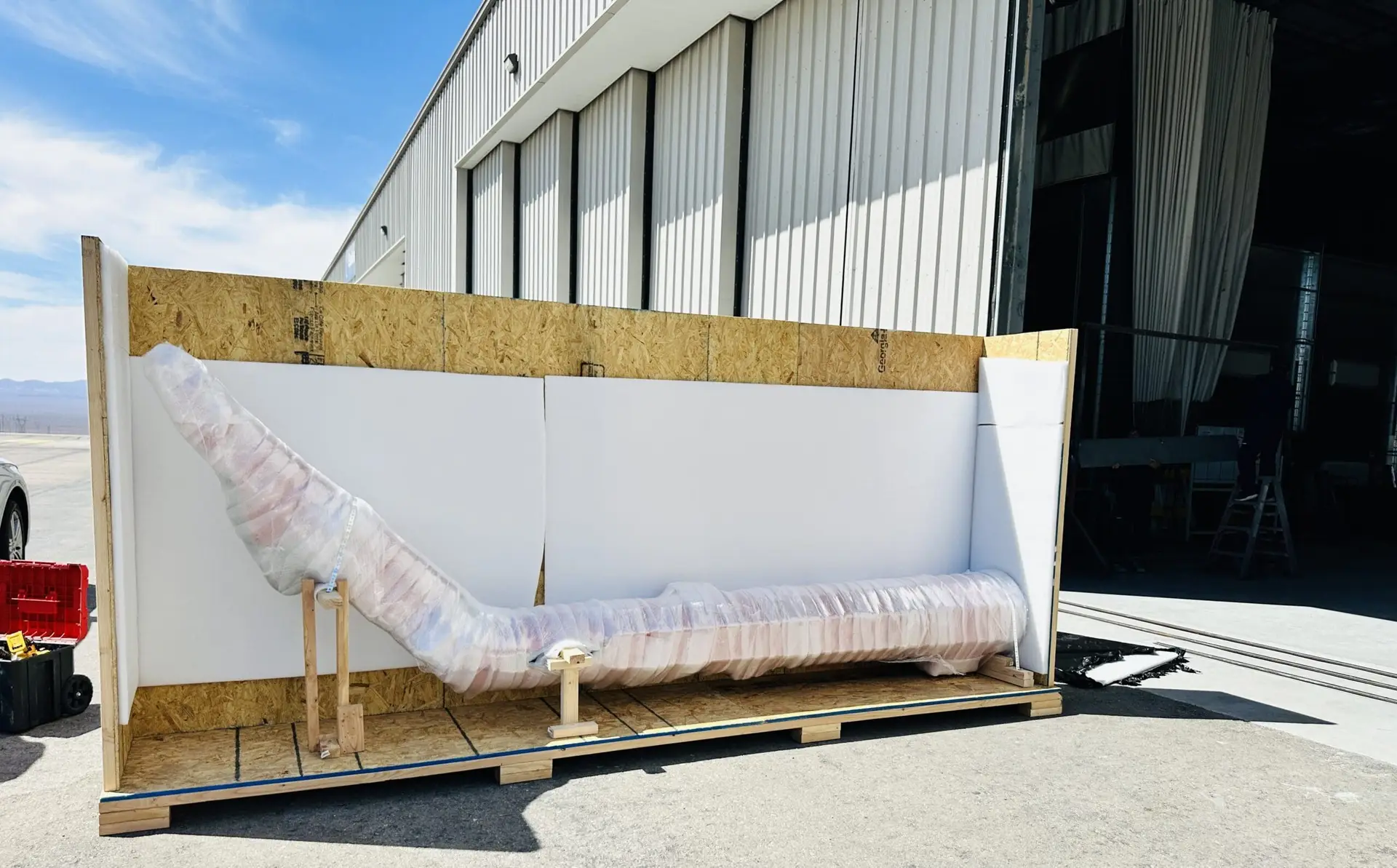 Wrapped curved object supported inside a large open wooden crate outdoors.
