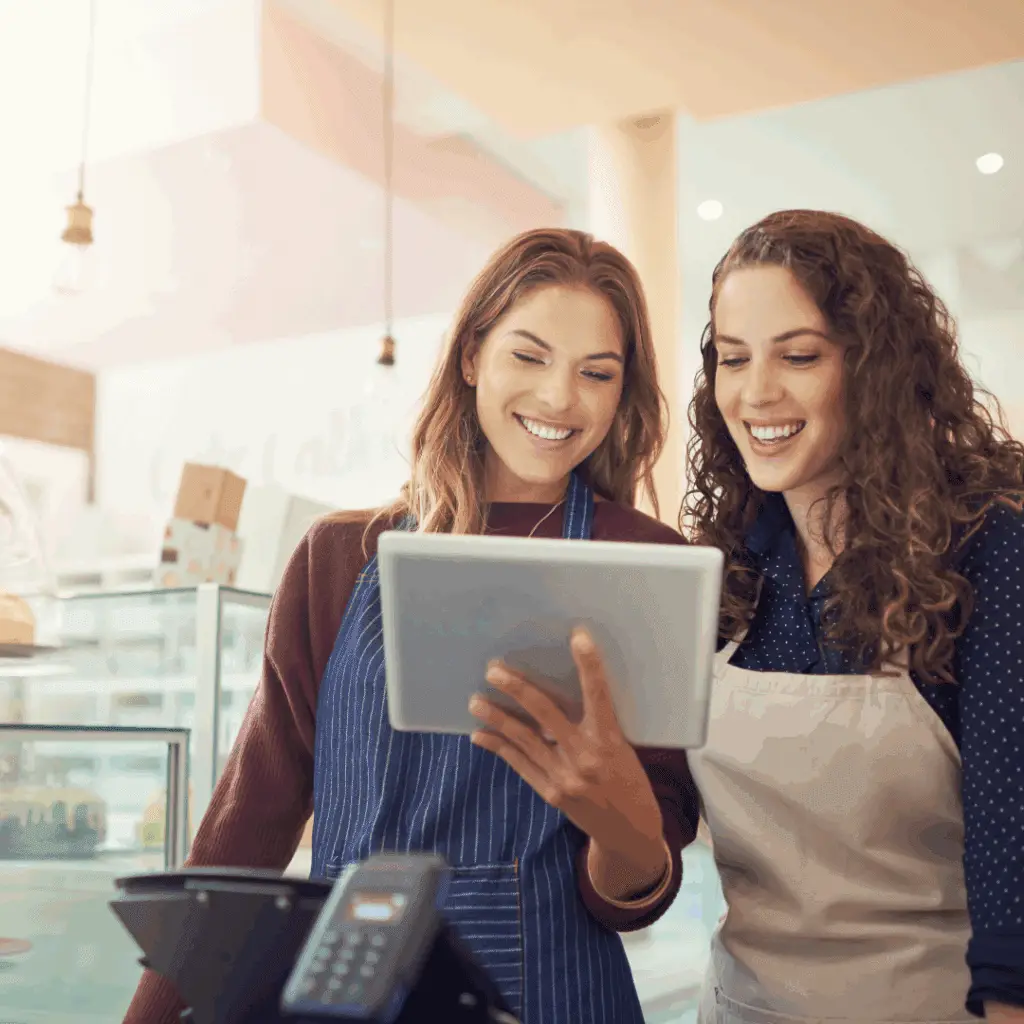 Two smiling women in aprons examining a tablet in a retail setting.