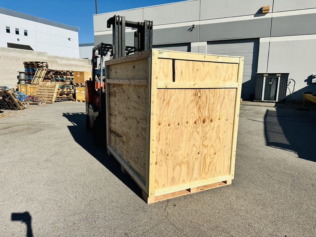 Forklift transporting a large, custom wooden crate in an industrial setting, surrounded by pallets and packaging materials, emphasizing heavy-duty crating solutions for secure shipping.
