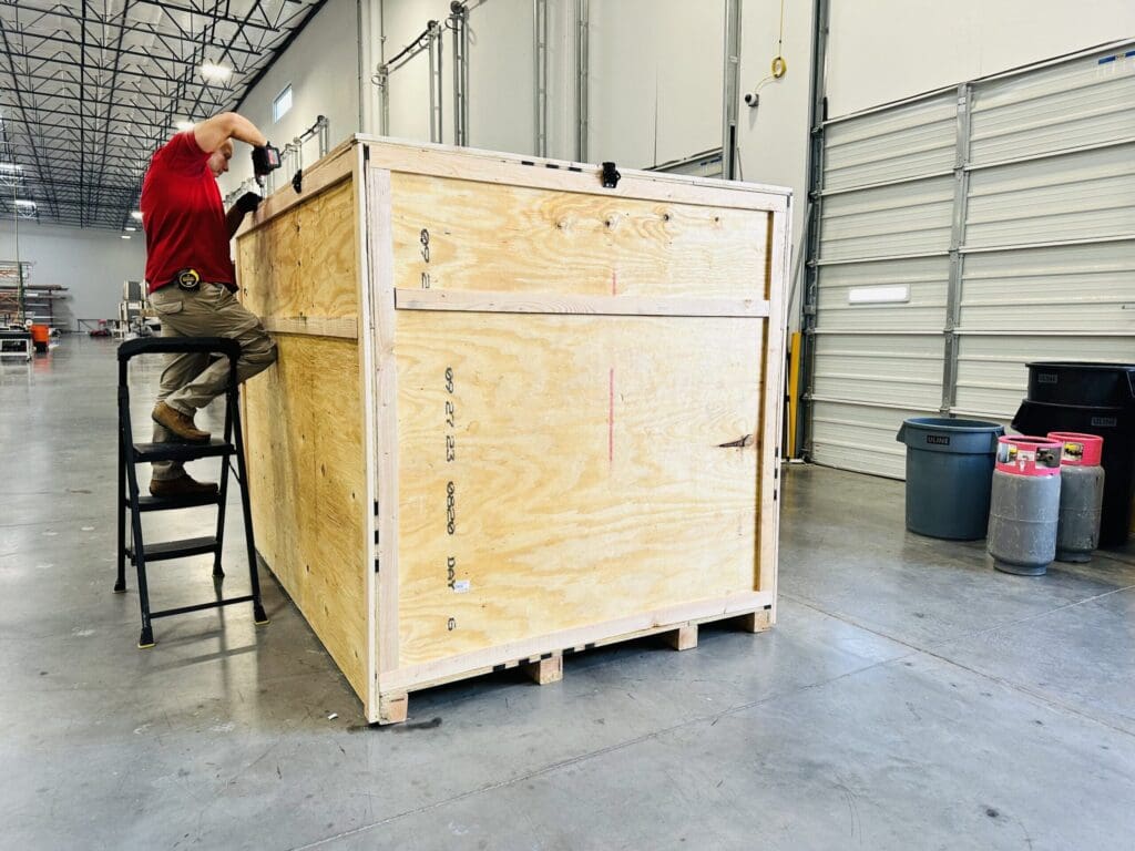 Worker assembling a custom wooden crate on-site, showcasing heavy-duty construction for industrial use in a warehouse setting.